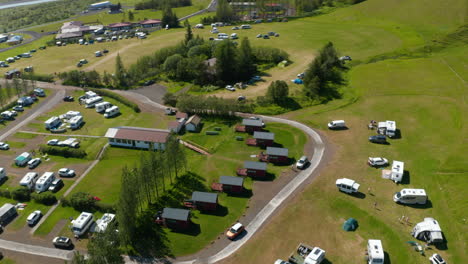 Direkt-über-Der-Luftaufnahme-Des-Campingplatzes-Im-Grünen-Wiesenhochland-In-Island.-Blick-Von-Oben-Auf-Den-Charakteristischen-Holzunterkunftsbungalow-Und-Touristencamping-Im-Wohnmobil