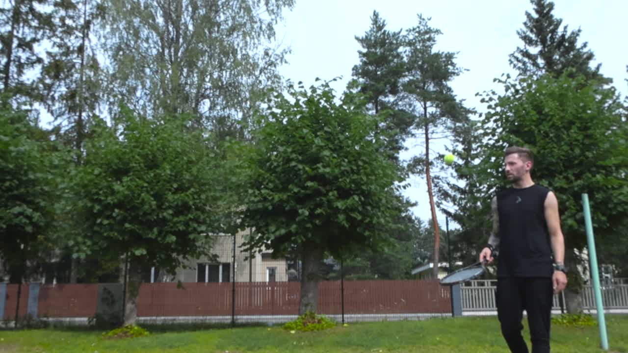 на кадре изображен татуированный мужчина, приближающийся к стене для тренировок в теннисе, собирающийся играть и практиковать теннис с яркими цветами в замедленном режиме и снят в разрешении 4k