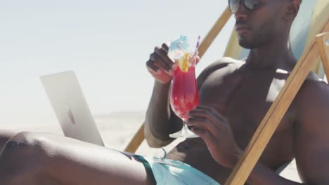 hombre afroamericano usando su computadora portátil en la playa y bebiendo jugo