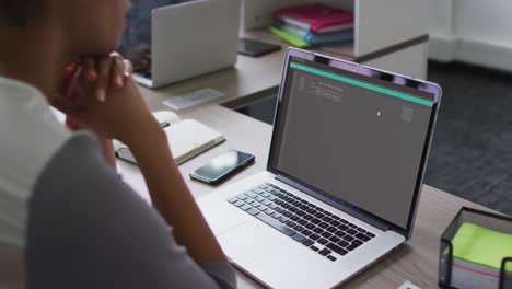 mujer de raza mixta sentada en un escritorio viendo el procesamiento de datos de codificación en la pantalla de un portátil