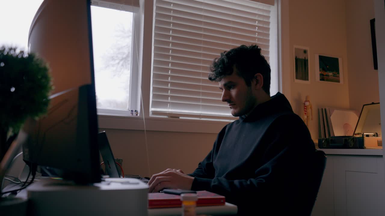 Primo piano di un uomo concentrato che lavora al computer in un ufficio domestico, con una tenue luce proveniente dalla finestra e un'illuminazione interna calda