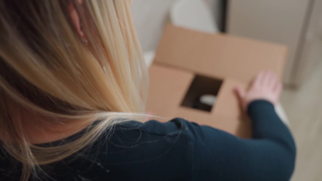 Vrouw pakt waterkoker in een doos, blonde vrouw zit aan een ronde tafel en plaatst waterkoker en mokken in een kartonnen doos, close-up van handen en dooskleppen, voorzichtige beweging, klein appartement interieur, verhuisdag.