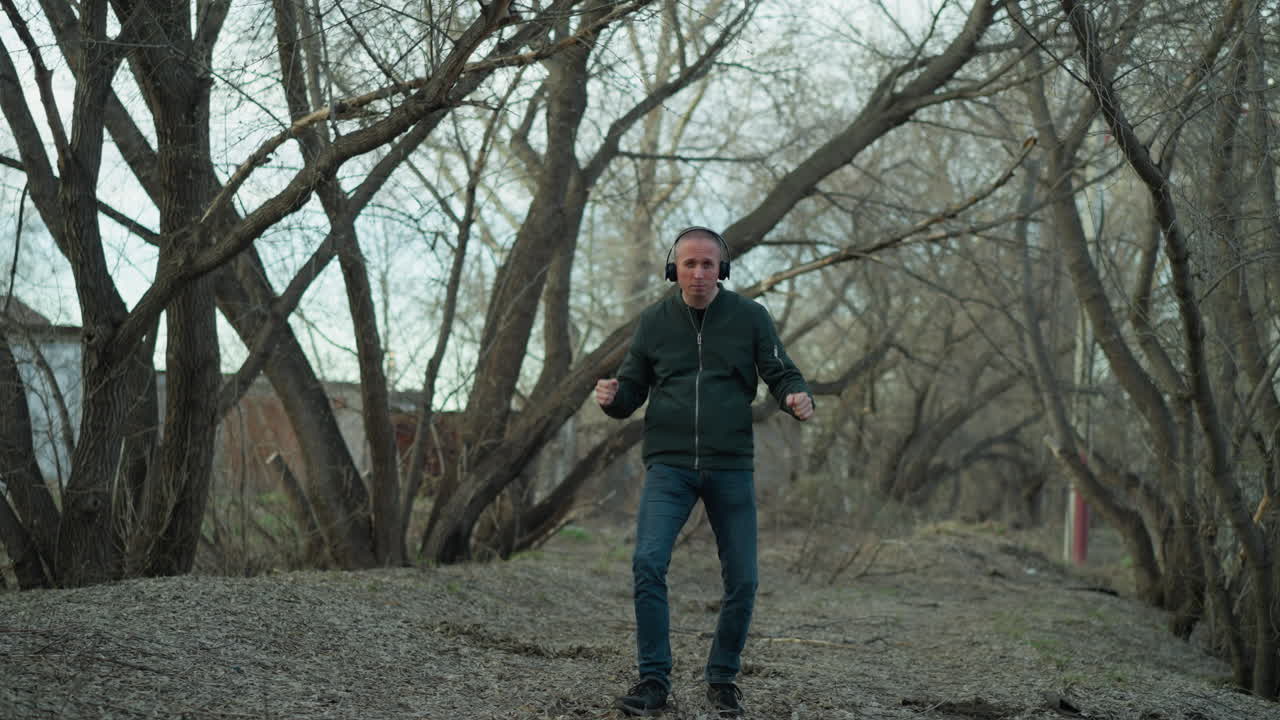 человек в зеленой куртке, с наушниками, весело танцует в спокойном лесу, он воплощает свободу и счастье в спокойной естественной обстановке, с деревьями и листьями вокруг него