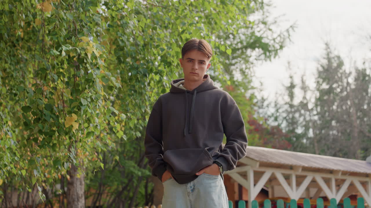 Adolescente caucasico con felpa con cappuccio, primo piano sotto il fogliame di betulla, sguardo pensieroso, mani in tasca, ambientazione nel cortile di periferia, luce soffusa autunnale, toni cinematografici suggestivi, i ruoli cambiano da studente