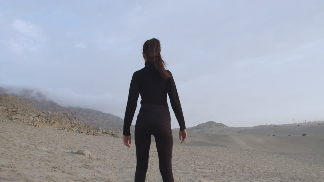 Woman from behind dressed in black with a strong pose standing in the middle of the desert with the cloudy sky and the wind blowing