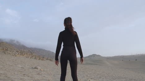 woman from behind dressed in black with a strong pose standing in the middle of the desert with the cloudy sky and the wind blowing