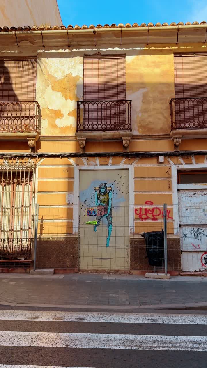 Alicante, España, 6 de agosto de 2025: Graffiti de arte callejero moderno en la fachada de un edificio antiguo. Mural urbano en una pared amarilla desgastada con la ilustración de un personaje debajo de balcones tradicionales.