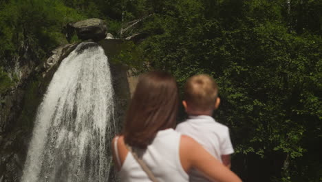 foamy waterfall sprays on steep cliff and mother with little son rests in wild park. woman shows whitewater cascade to toddler boy on weekend hike