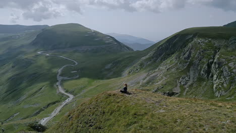 individual sits peacefully on a grassy edge, gazing at the vast, winding landscapes of the transalpina in romania, under a partly cloudy sky