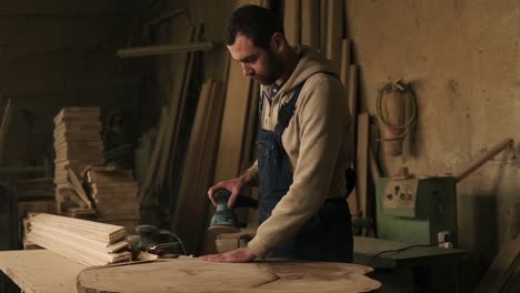 Eine-Tischlerei-Mit-Holzmaterial,-Werkzeuge-Im-Hintergrund.-Tischler,-Der-In-Blauen-Overalls-An-Der-Holzverarbeitung-Arbeitet.-Schleift-Das-Material-Sorgfältig-Mit-Einer-Handmaschine