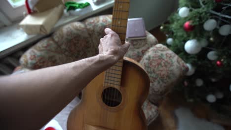 close-up old senior in traditional red santa claus costume with white beard sits near decorated christmas tree, plays the guitar, concept of congratulations, merry christmas.