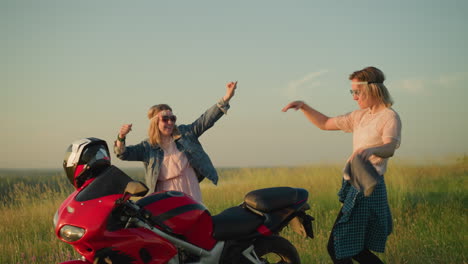 two joyful women are dancing near a red motorcycle in an open field as one of them tosses a cloth to her friend, who catches it and continues dancing with excitement