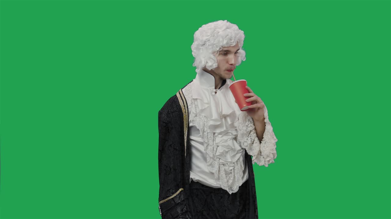Portrait of courtier gentleman in vintage suit and wig, drinking drink from plastic glass through straw. Young man posing in studio with green screen background. Close up. Slow motion ready 59.94fps