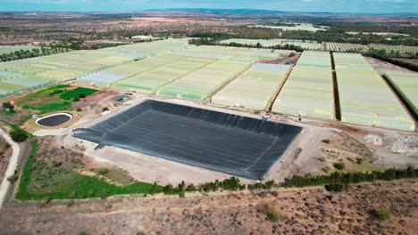 Vista-Aérea-De-Invernaderos-En-Campo-Agrícola-Con-Cuenca-Vacía-Para-Suministro-De-Agua.