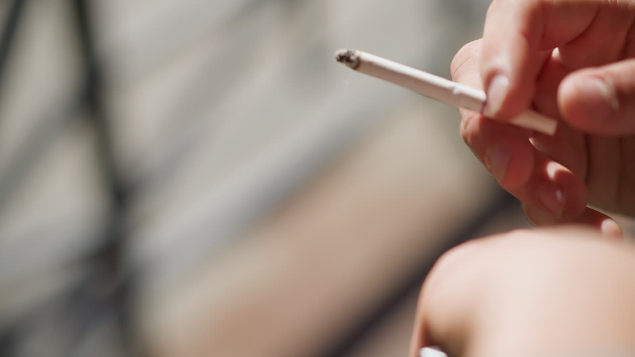 close up hand view of white man holding cigarette between fingers while smoking in natural light with soft blur background showing gentle curls of smoke and warm tones of outdoor setting