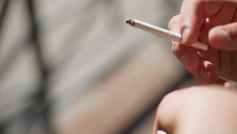 close up hand view of white man holding cigarette between fingers while smoking in natural light with soft blur background showing gentle curls of smoke and warm tones of outdoor setting