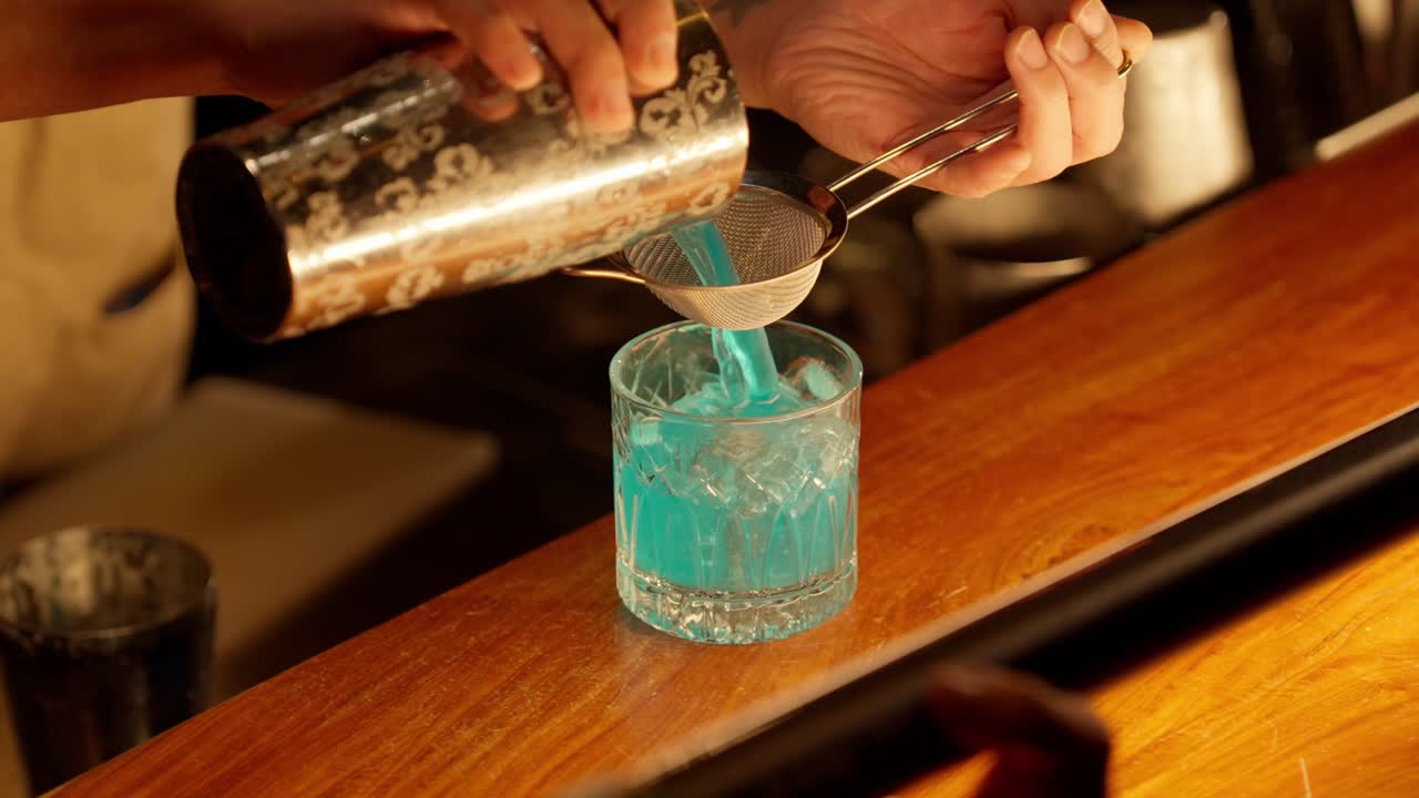 Um bartender profissional usa uma coqueteleira Hawthorne e um coador de malha fina para coar duplamente um vibrante coquetel azul em um copo rocks de cristal cheio de gelo em um bar badalado.