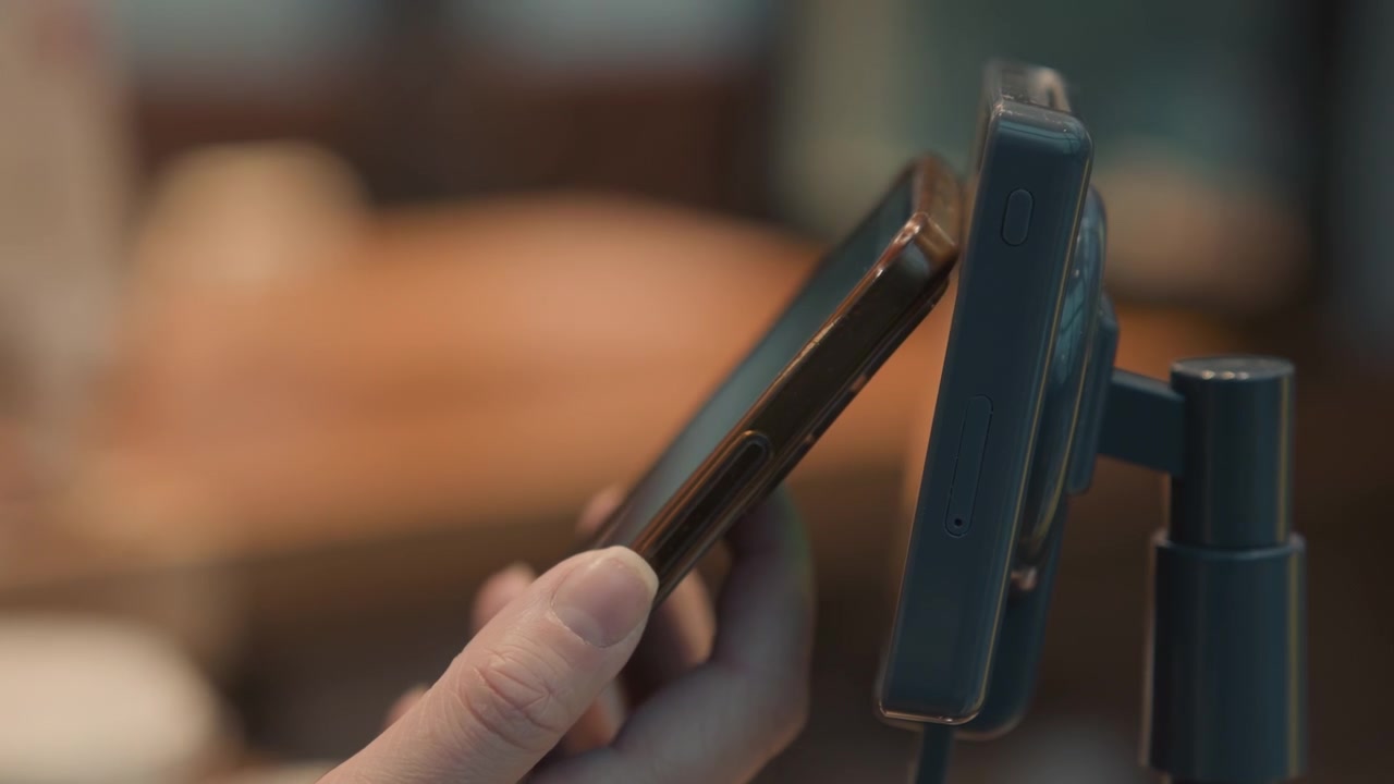 Smartphone tap on card reader terminal, hand holding phone performing contactless payment at cozy cafe counter with blurred bokeh background, mounted reader on adjustable stand, warm lighting