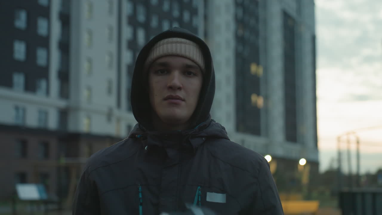 hombre con capucha y gorra realizando control de pelota con una pelota de fútbol durante una sesión de entrenamiento urbano, mostrando enfoque y habilidad contra el telón de fondo de altos edificios residenciales y cielo nublado por la noche