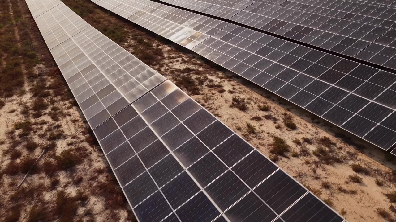 Riprese aeree mostrano file di pannelli solari che luccicano sotto il sole estivo, catturando energia rinnovabile in un vasto impianto solare spagnolo, con terra arida e vegetazione naturale sottostante.