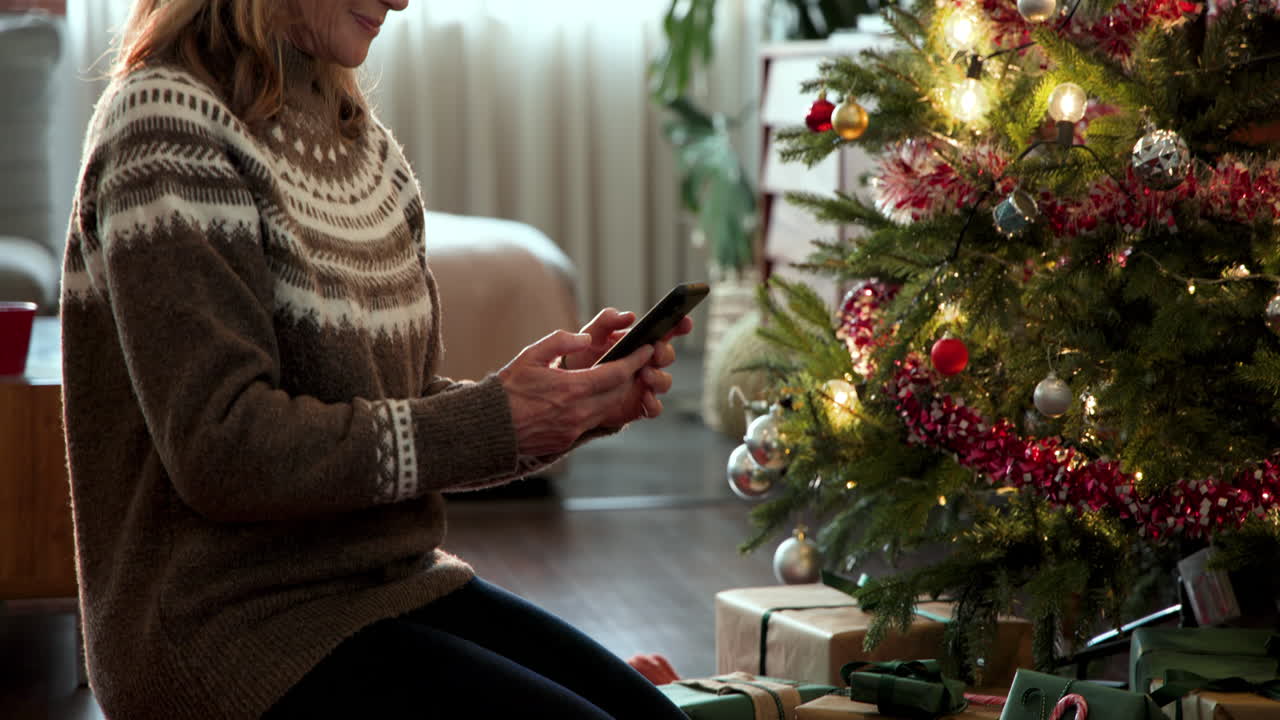 Senior woman using smartphone near decorated Christmas tree at home, feeling festive