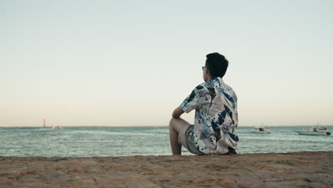 Hombre-Asiático-Sentado-En-Una-Roca-Hacia-La-Playa-Relajarse-En-Vacaciones-Con-Gafas-De-Sol-Mirando-Hacia-Atrás