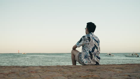 asian man sitting on rock toward beach relax on vacation holiday with sunglasses looking back