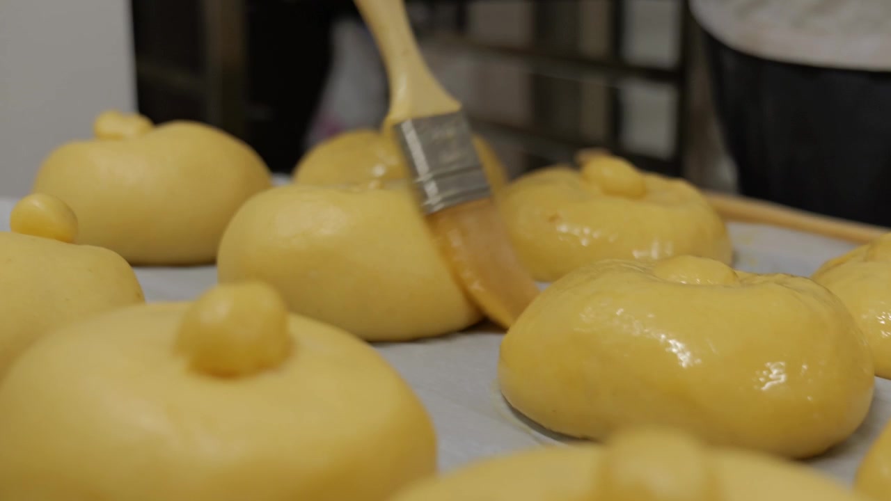 Padeiro passa um pincel com ovo batido sobre várias brioches parisienses enfileiradas