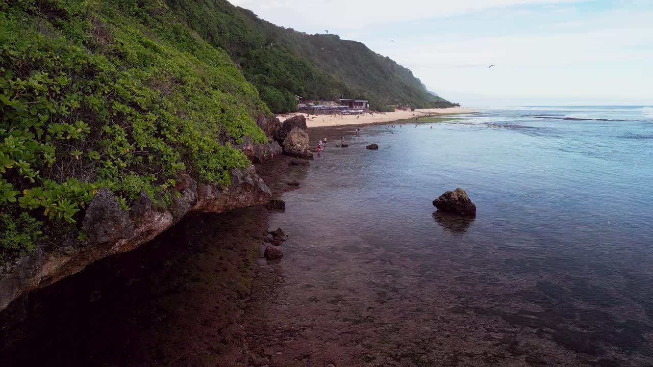 Soaring drone angle shows a long Bali beach during low tide with smooth water patterns meeting rugged shoreline rock while a towering coastal green hill forms a dramatic backdrop for relaxed visitors
