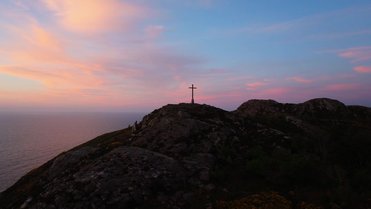 La prima luce del giorno proietta un tenue bagliore su una croce in cima a una collina, con nuvole rosa che fluttuano sopra il mare. Adatto per temi spirituali o contenuti di viaggio
