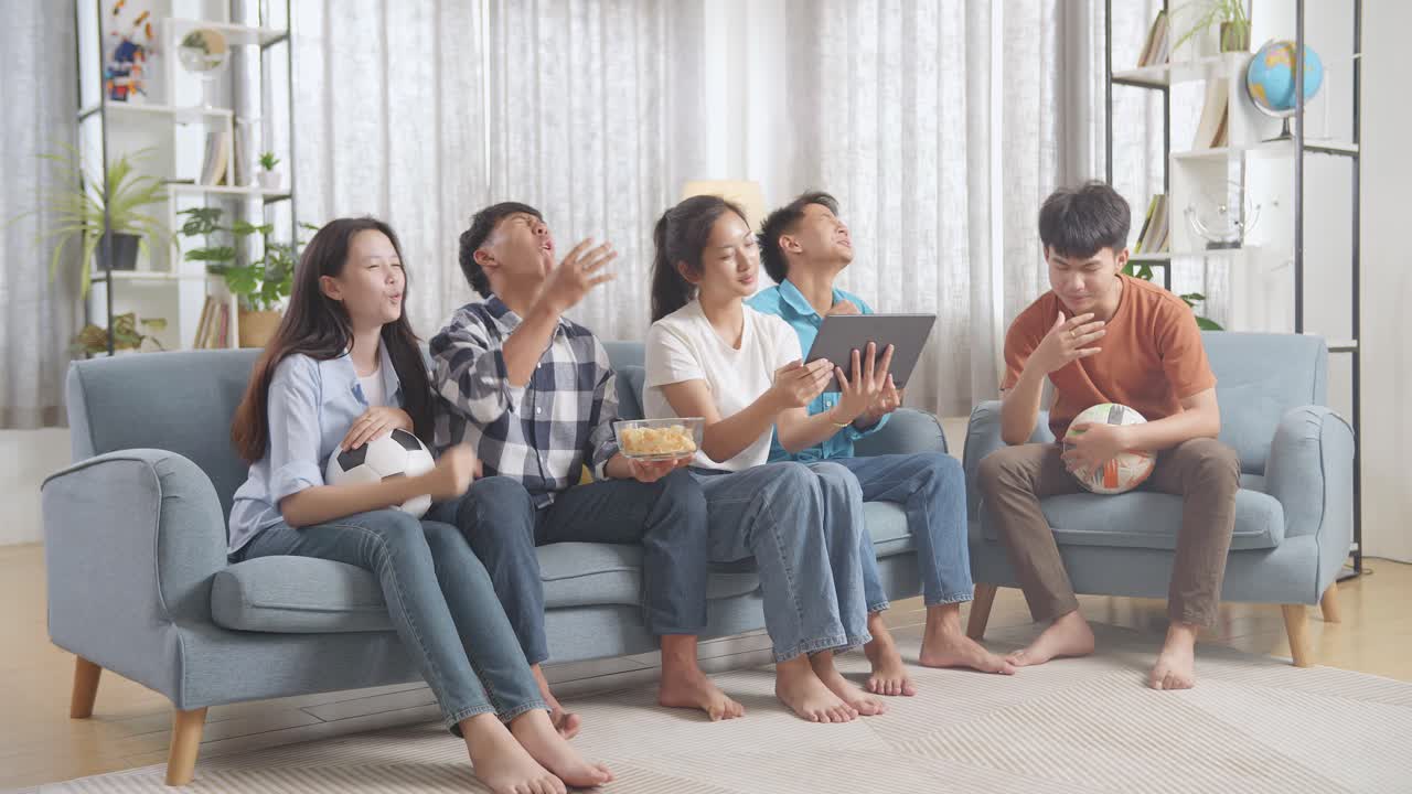 아시아 십대들이 태블릿에서 축구 경기를 응원하고 지켜보고 집에서 실망합니다.