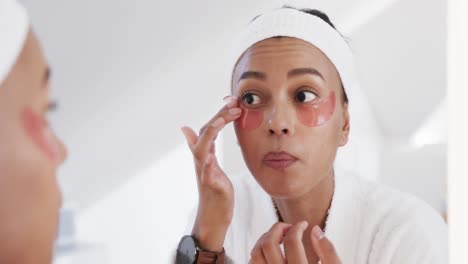 Focused-biracial-woman-applying-under-eye-masks-looking-in-mirror-in-sunny-bathroom,-slow-motion