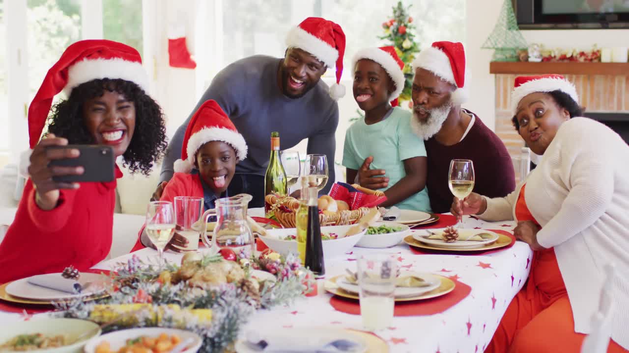 Famiglia afroamericana con cappelli di Babbo Natale che si scatta un selfie con lo smartphone mentre è seduta a tavola.