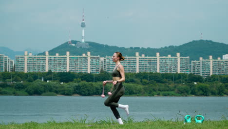 Athletische-Hübsche-Frau-In-20.-Übungen-Mit-Kabellosem-Springseil,-Die-Auf-Einem-Bein-Und-Beiden-Beinen-Auf-Einer-Grasbewachsenen-Flussuferwiese-Im-Han-River-Park-Mit-Dem-Seoul-Namsan-Tower-Dahinter-Springt-–-Profilansicht-In-Zeitlupe