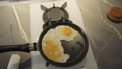 close-up shot of frying pan with two eggs cooking in modern kitchen, yolks intact, sizzling on stove top, with kitchen utensils and countertop visible, focused on food preparation process