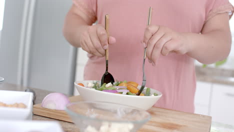 midsection of plus size biracial woman tossing bowl of feta salad in kitchen, slow motion