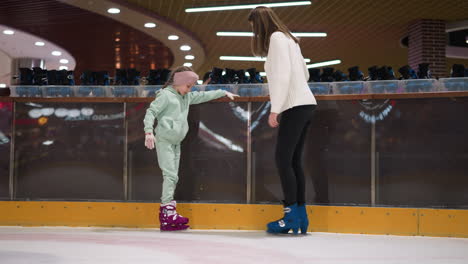 a young girl in a mint green outfit and pink skates is standing on an ice rink, holding onto the edge, and blue skates stands beside her, with rows of rental skates in the background