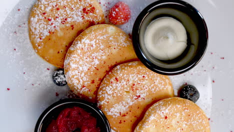 eastern european cuisine offers small pancakes served with powdered sugar, sour cream, berry jam, raspberries and blueberries on a white plate and a glass of orange juice on a table