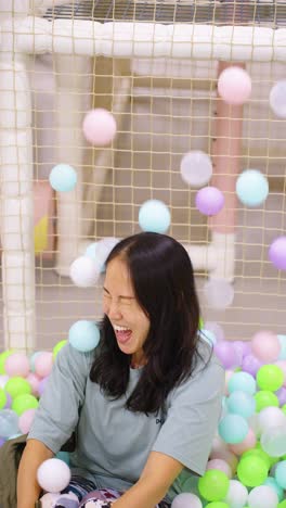 a woman joyfully laughs as colorful balls fall around her in a playful indoor ball pit
