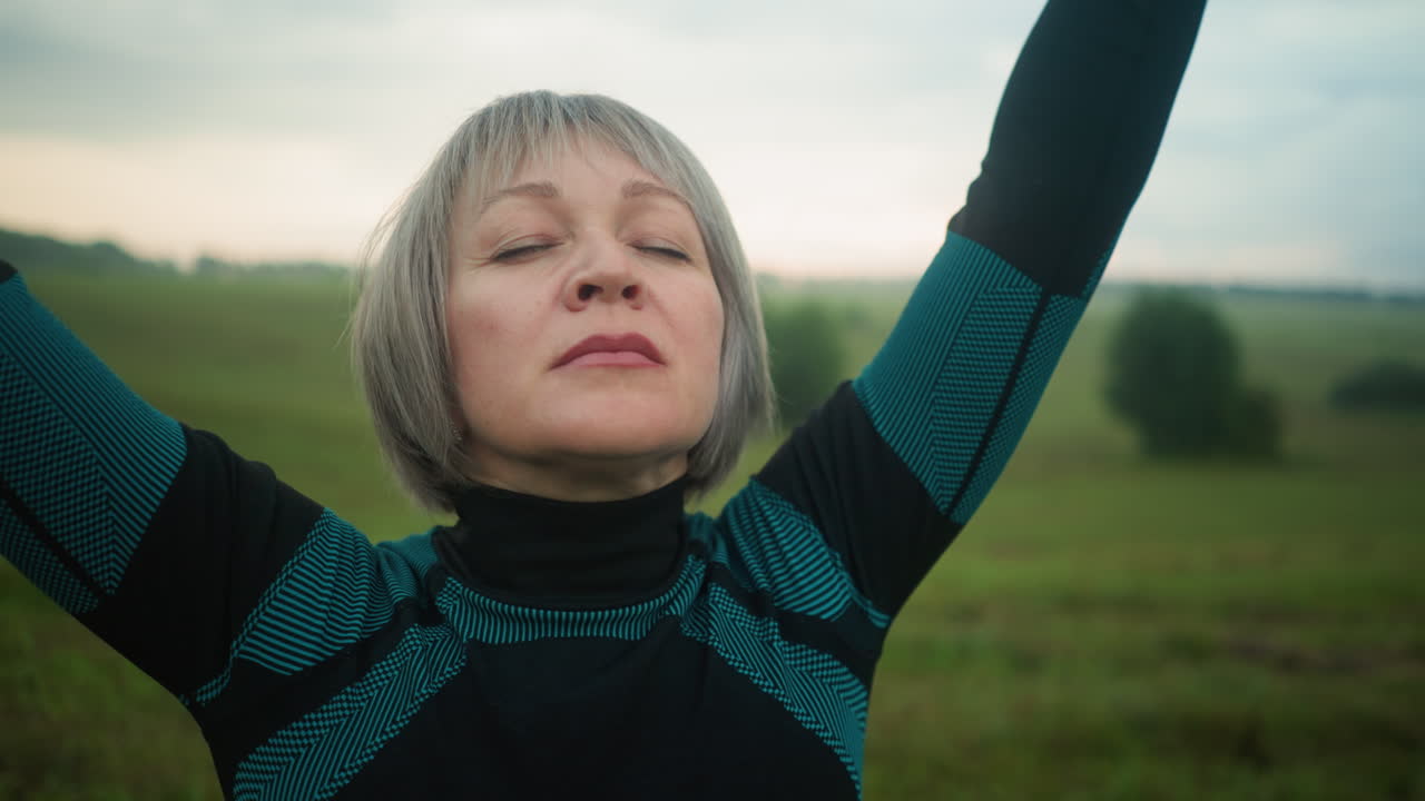 Close-up of white woman standing under cloudy sky with eyes closed, practicing yoga, she slowly lifts her arms and lowers them, focusing on mindfulness, relaxation, and deep breathing