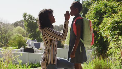 african american mother putting on face mask on daughter with backpack outdoors on a bright sunny da