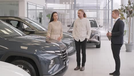 young car salesman showing to young couple new automobile at dealership salon.