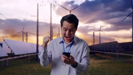 asian male professional worker standing with his smartphone with solar panel and wind turbines, he raises his fist up with screaming goal