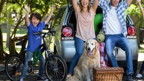 happy caucasian parents, son and daughter by car cheering, with golden retriever pet dog in park