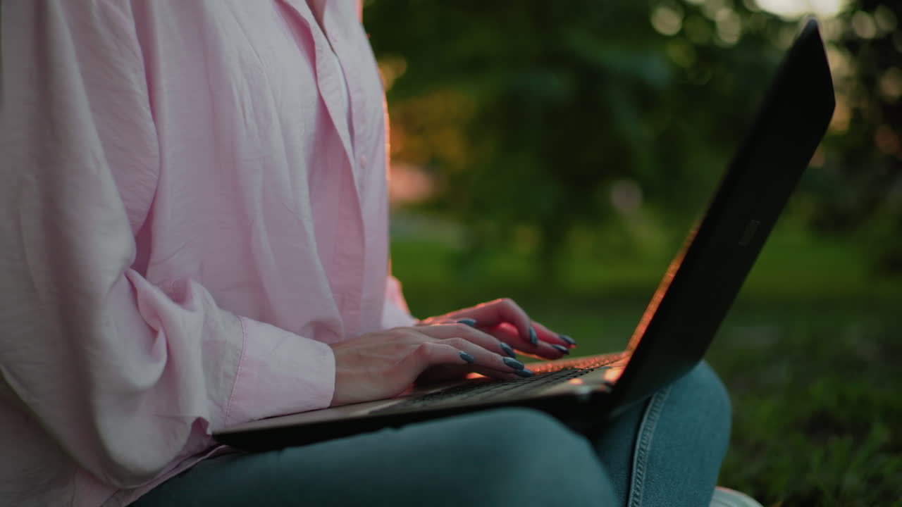 Close-up of remote worker in pink shirt with polished nails typing on her laptop, bokeh light effect reflecting off her hands with flies flying around, and background features lush greenery