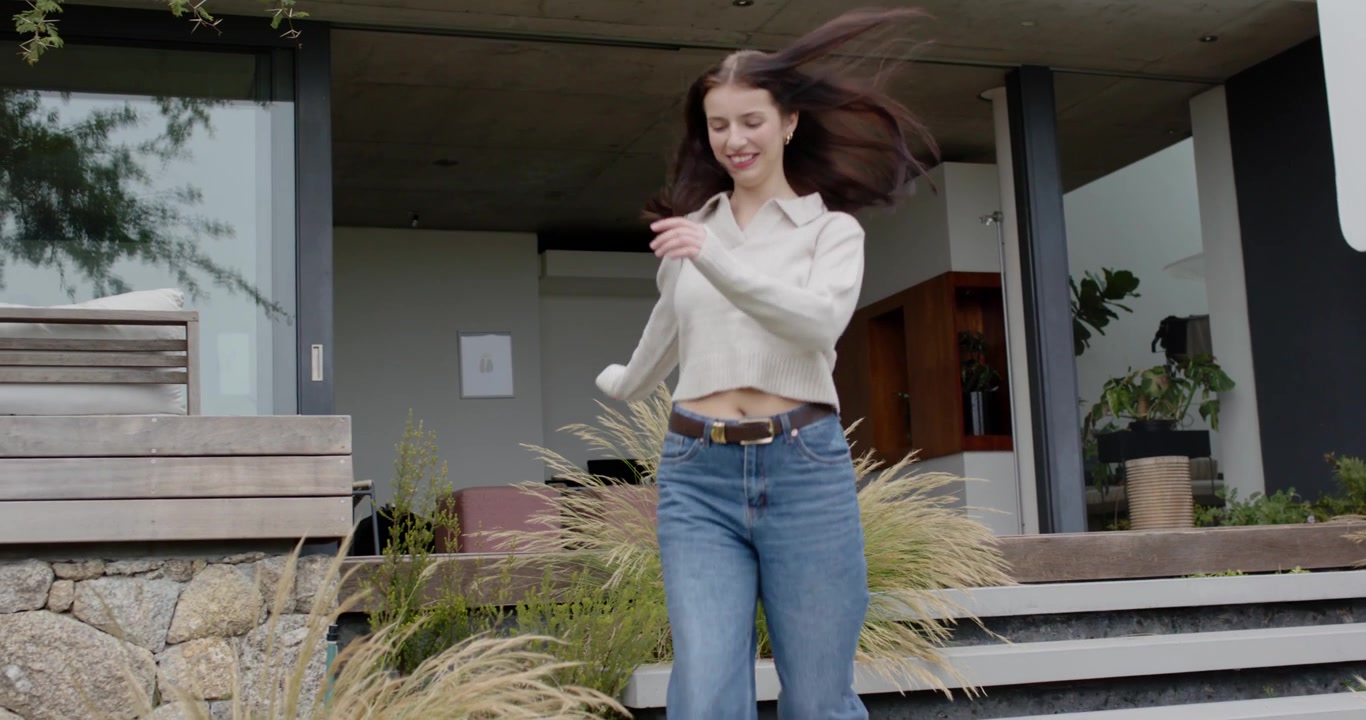 Voksen kvinde i sweater og jeans går ud fra overdækket veranda, går ned ad trappen, græs børster linsen.