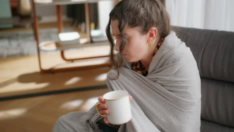 woman with cold sits wrapped in blanket and drinks tea in living room