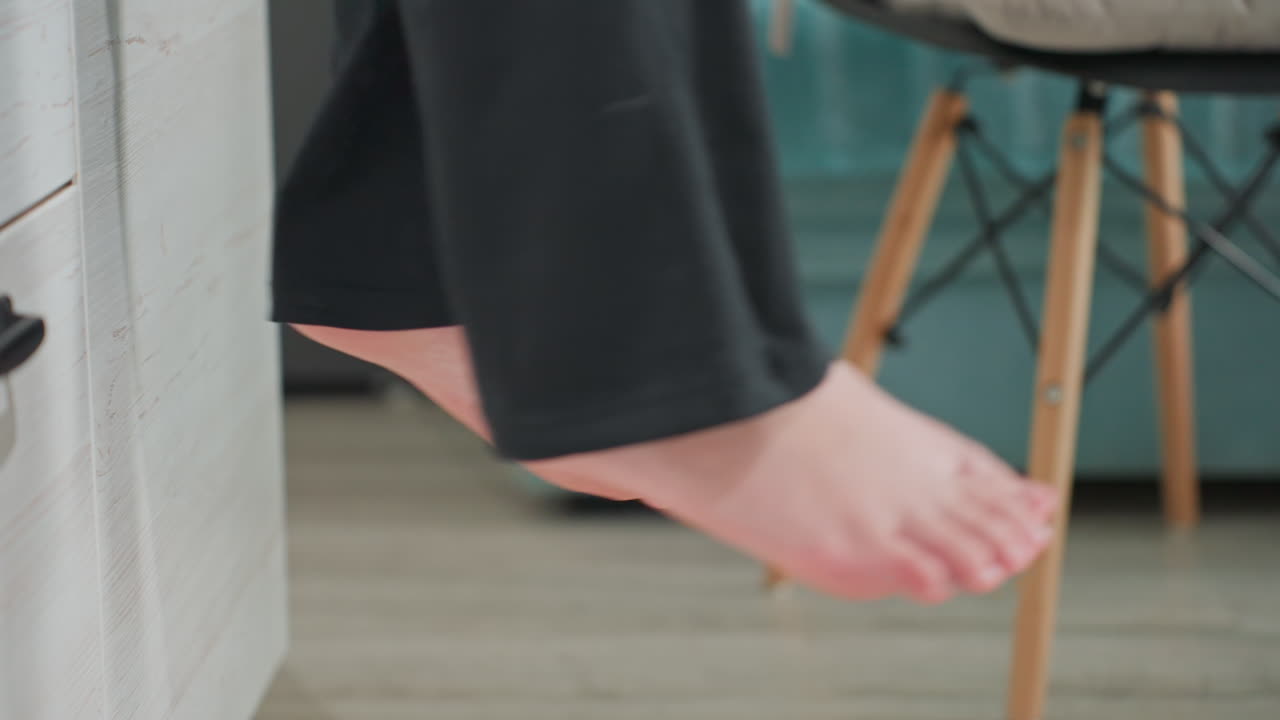 side view of adult legs swinging while seated on white cabinet edge inside bright modern kitchen setting with soft natural light highlighting smooth movement of black pants contrasting pale wood