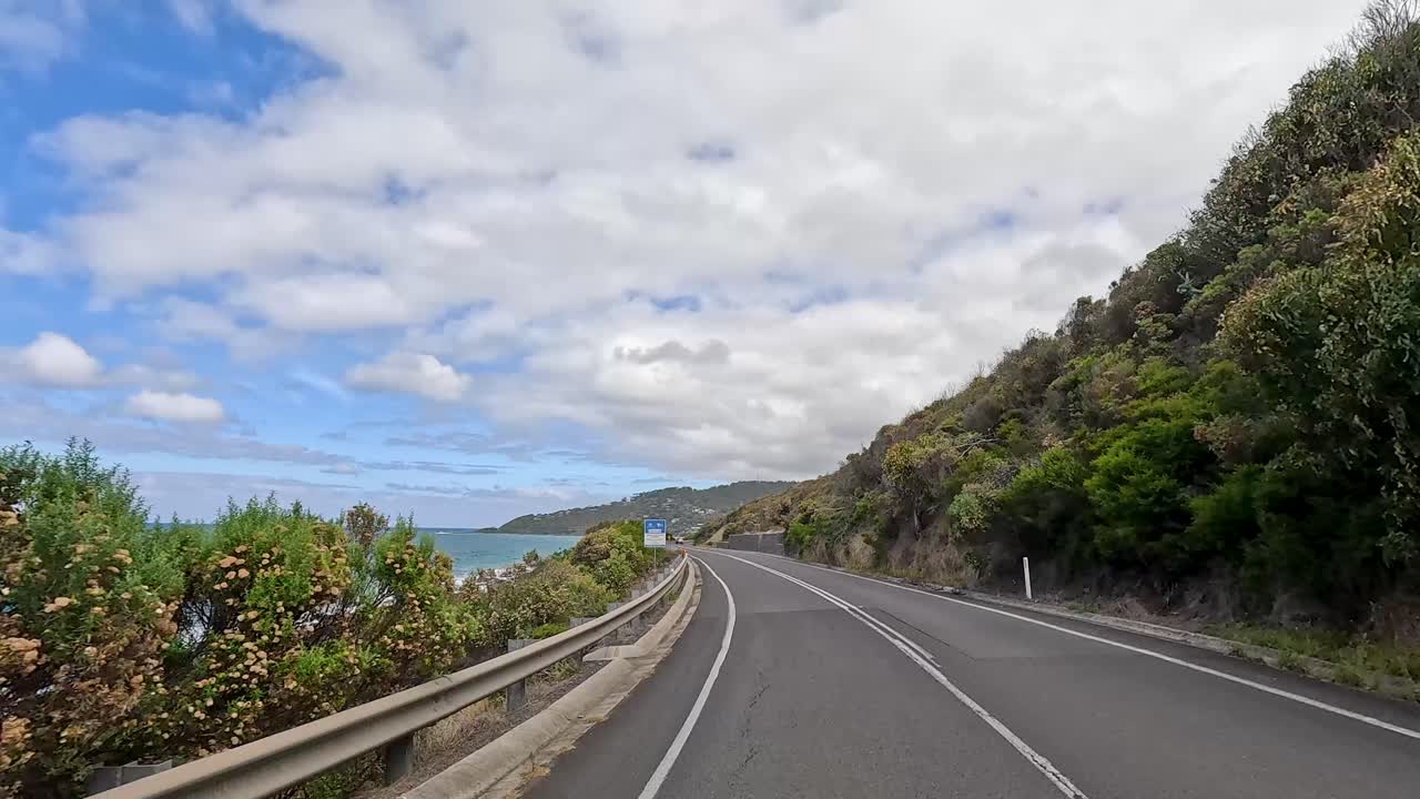 A 14-second drive showcasing lush greenery and ocean views along the Great Ocean Road under a partly cloudy sky