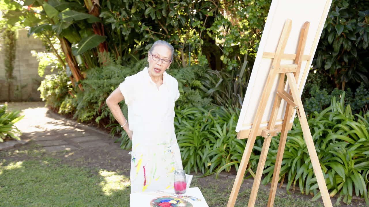 Painting on canvas, elderly woman standing in garden with easel and paint supplies